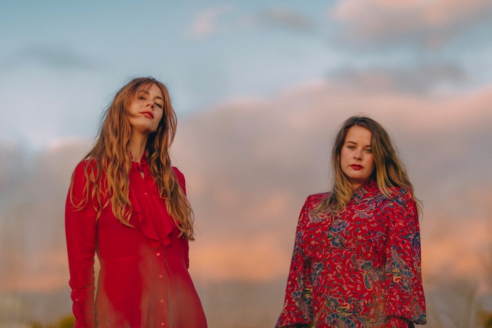 A photograph of the duo Dolores Forever, both wearing red summery dresses and dark blonde hair with a sunset as the backdrop.