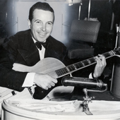 Nappy Lamare holding a guitar and sitting behind a desk with music while looking forward and smiling.png