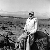 Ernest Hemingway sits on a large rock while holding a rifle in Africa.jpg